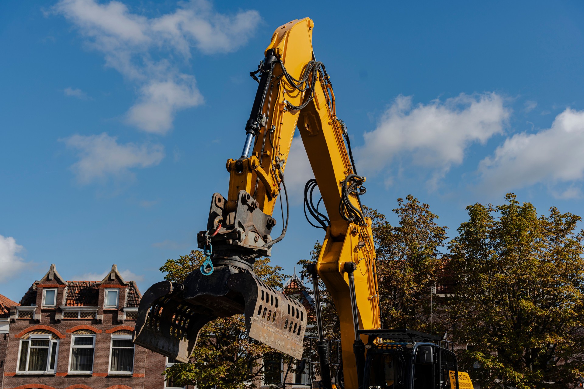 Yellow excavator reaches toward the sky, towering above houses and trees on a partly cloudy day.